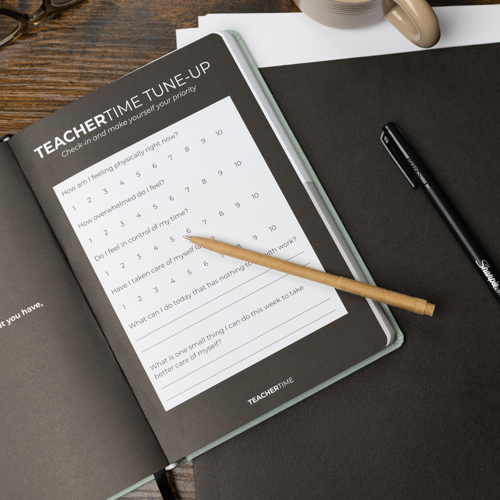 A TEACHERTIME 'Tune-Up' planner page shown in a styled flat-lay on a dark desk with a wooden surface, a black pen, and a pencil.